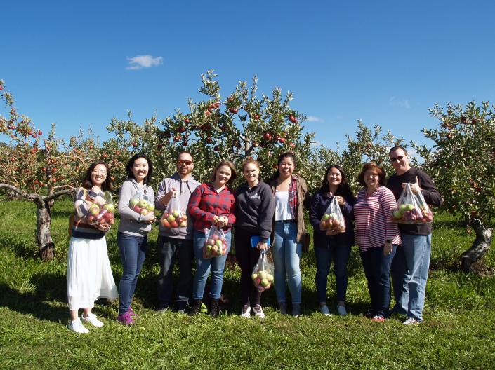 Image of 9 people standing in front of apple trees and holding bags of apples