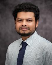 Headshot of Atikur Rahman in a blue and green thin lined striped shirt with a blue tie and grey blurred background