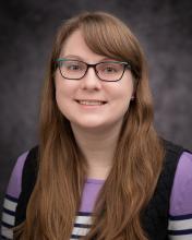 Headshot of Brianna Bauer in a purple black and white striped shirt and black vest with a blurred grey background