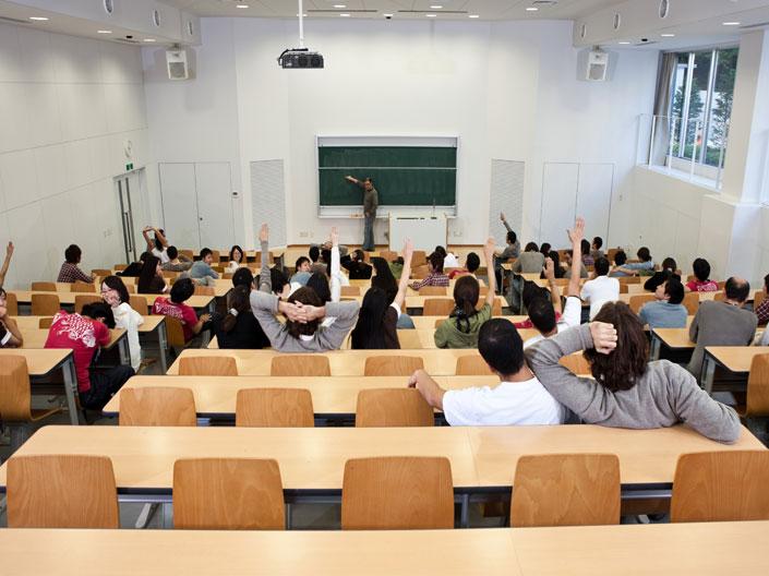 View from the back of a lecture hall filled with students facing the front of a room, where a faculty member stands talking to the students. 