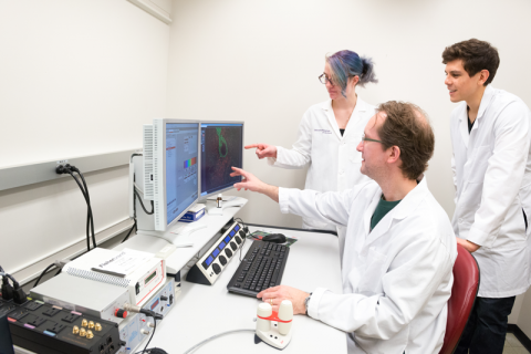 Two young researchers in white lab coats stand with Dr. Brian Cobb in white coat to analyze data on a computer screen.