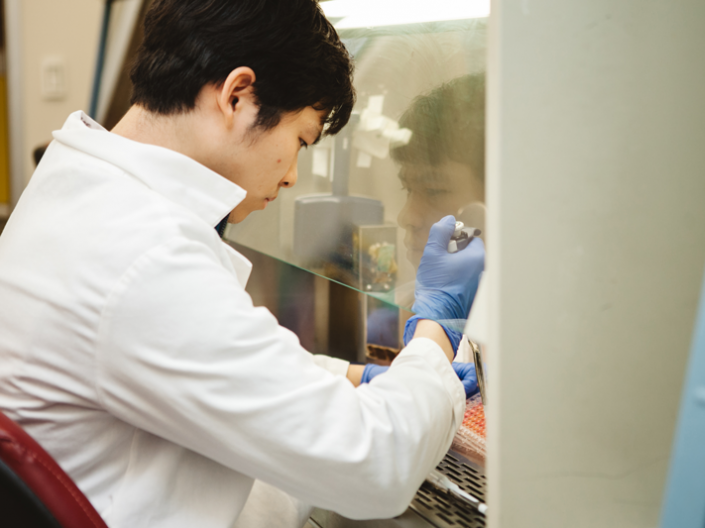 Man in white coat working with a pipette under a clear laboratory fume hood