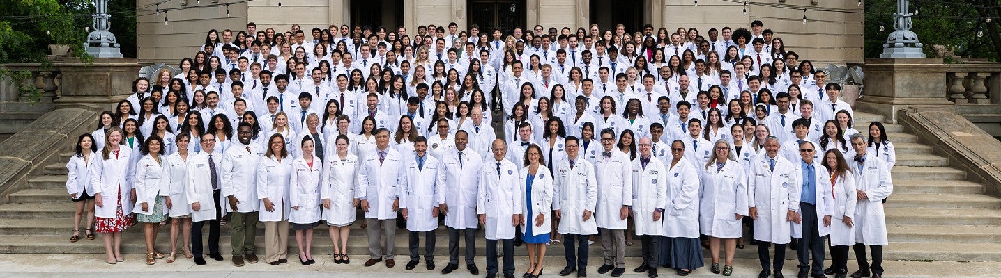 A large group of physicians in white coats stand on the stairs of the Maltz Center and pose for a photo.