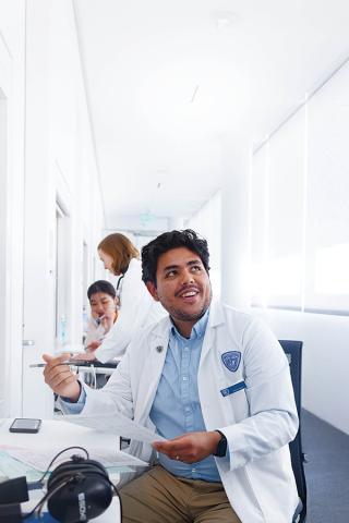 A School of Medicine student in a white coat sits and smiles while looking up to the right corner of the frame.