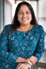Sudha wearing a blue patterned shirt sitting with her hands crossed 