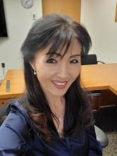 Headshot of Dr. Sua Myong; individual with long dark hair and bangs, dark blue shirt, office desk in the background. 