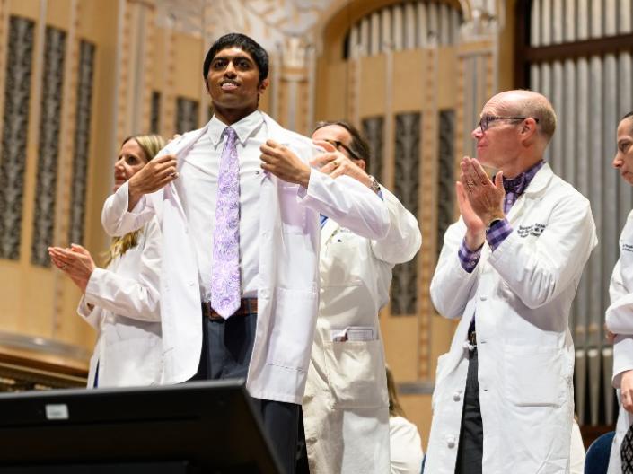 A student gets their white coat on stage while doctors clap around him.