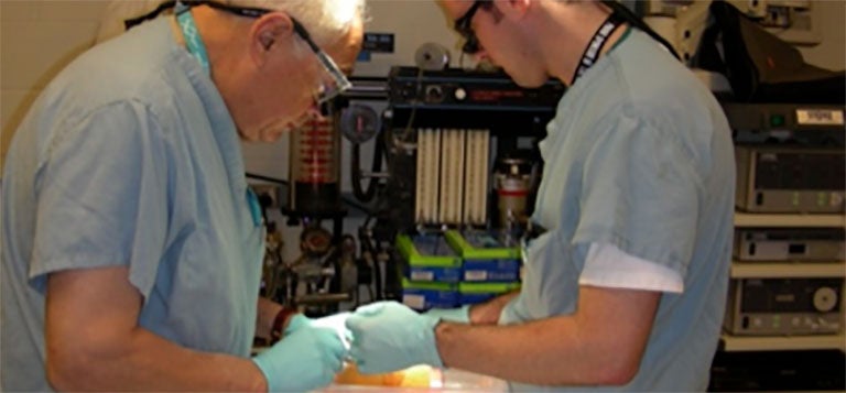 two surgeons working in a room with equipment and supplies against the far wall