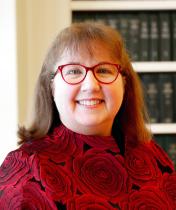 Portrait of Tammy Randall wearing a red shirt and red glasses sitting in front of a book case.