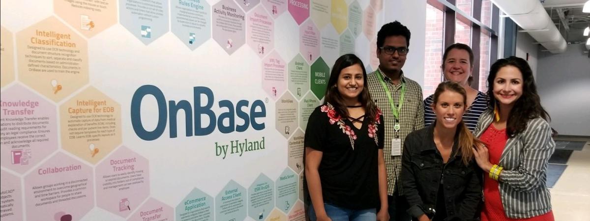 An adviser and a group of students standing in front of OnBase logo indoors  