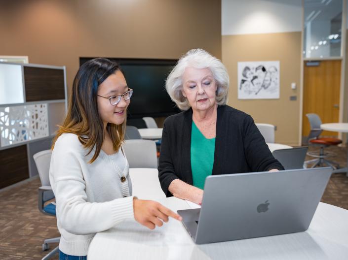 Grey-haired woman helping dark-haired woman at a laptop in a modern setting