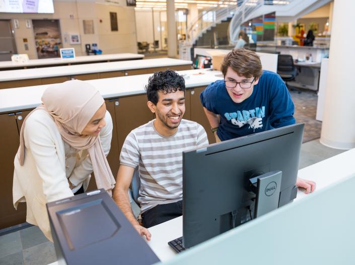 Three multi-ethnic students smiling around a computer together