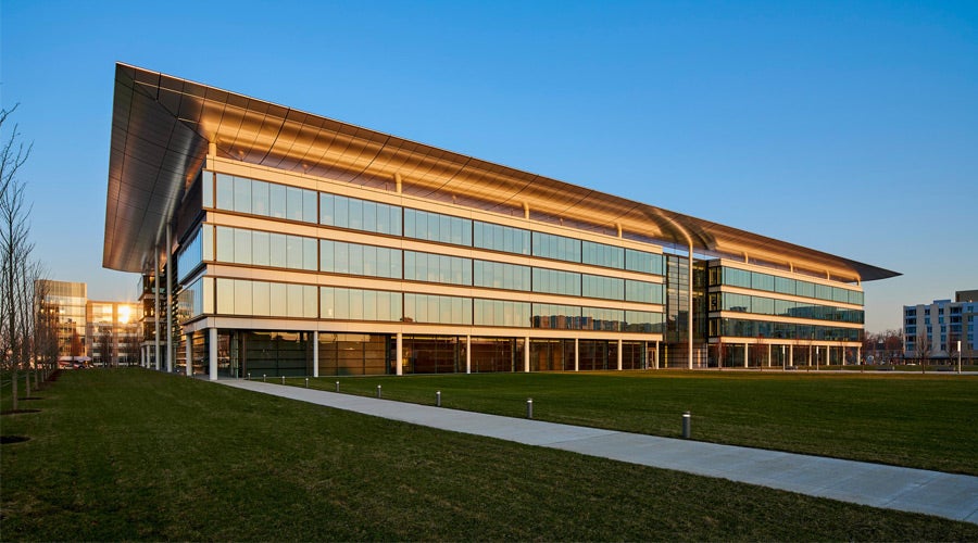 Modern building with large glass windows under a clear blue sky, reflecting a golden sunset. It is surrounded by a vast green lawn and walkway.