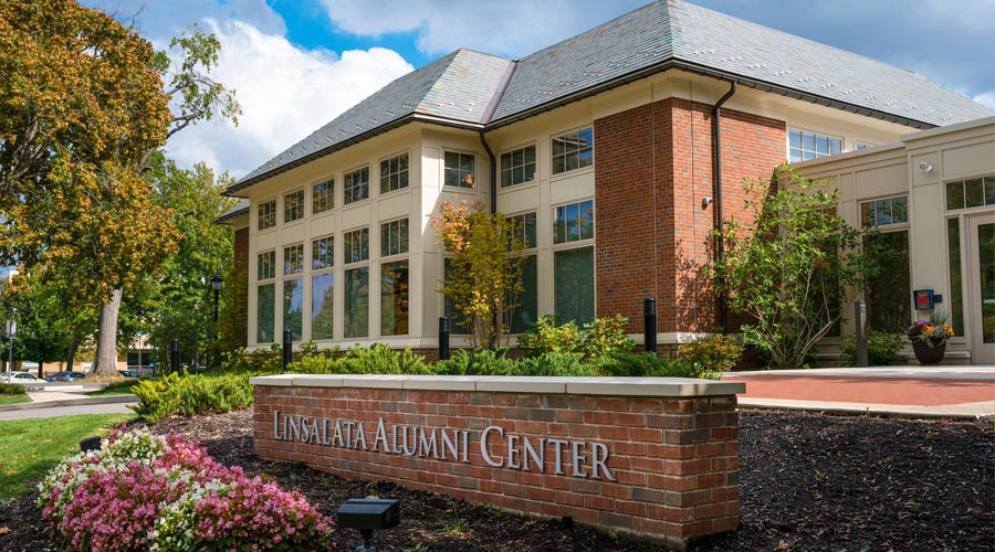 Red-brick building with large windows, surrounded by lush greenery and vibrant flowers under a partly cloudy sky. Sign reads “Linsalata Alumni Center.”