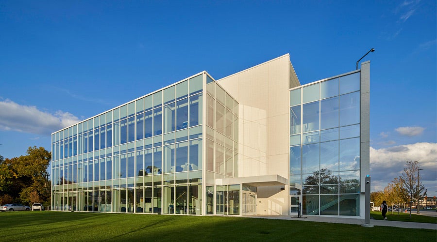A modern, glass-fronted building with three stories reflecting clear blue skies, set against a backdrop of trees. The scene conveys a crisp, professional atmosphere.