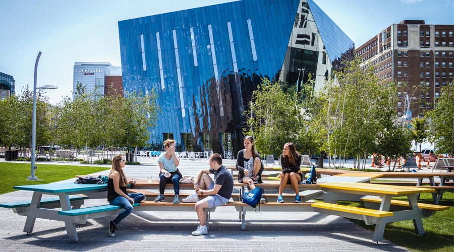 A group of young adults sits casually on colorful outdoor benches in front of a modern, angular glass building, surrounded by trees, on a sunny day.