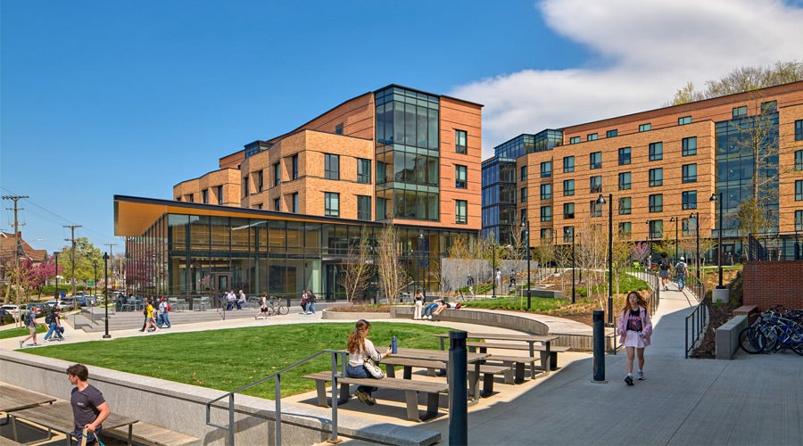 Modern university campus with brick buildings under a blue sky. Students walk and sit on benches in a landscaped courtyard, creating a lively atmosphere.