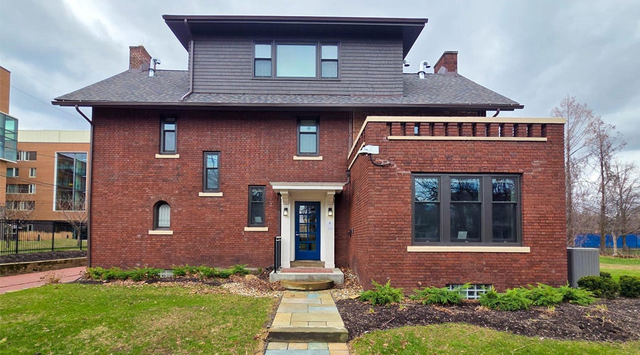 A two-story brick house with a modern design, featuring a prominent blue door, small front porch, and multiple narrow windows. The yard is neatly landscaped with grass and shrubs.