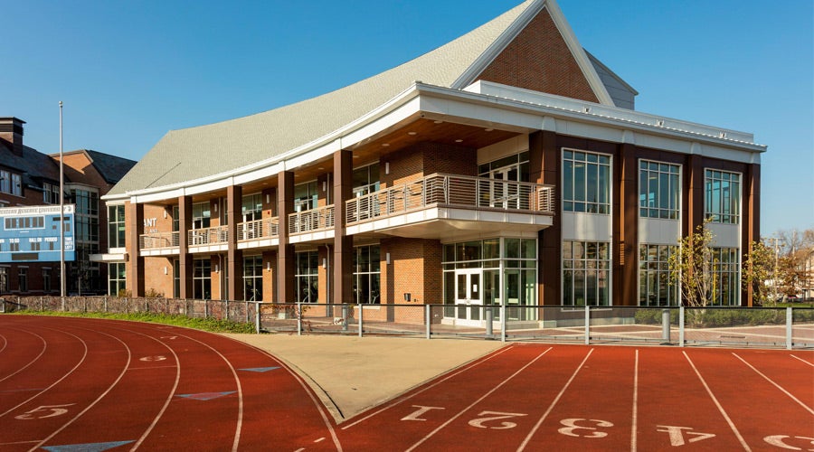 Modern building with a sloped roof, large windows, and balconies, set against a clear blue sky. Red running track curves in the foreground.