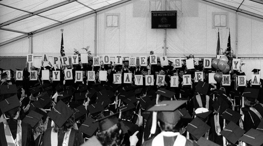Black and white image of graduates on stage in a tent on campus holding letter cards that spell out "Happy Mother's Day from Your Favorite MBA"