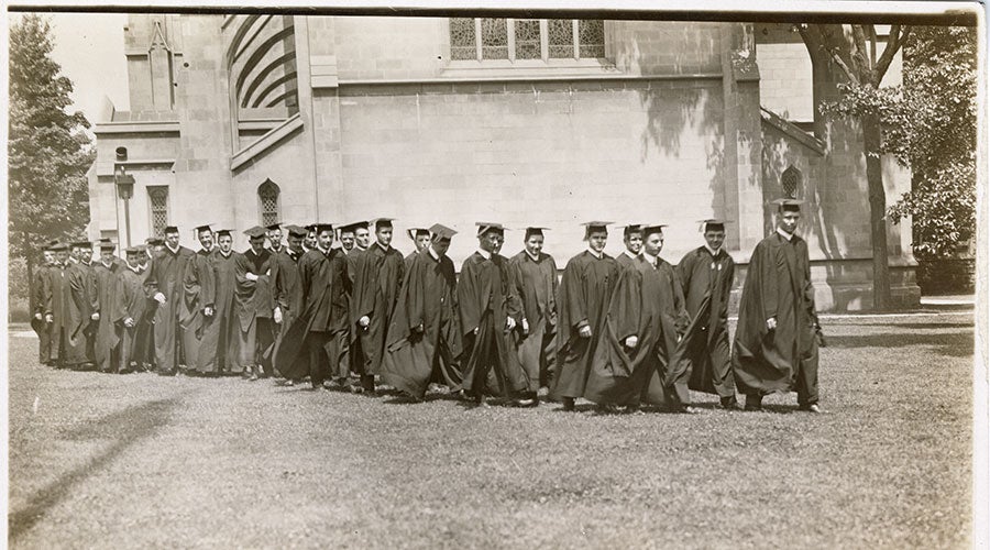 A black and white photo of men wearing academic robes walking in a line on campus