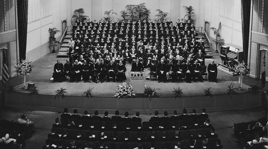 Black and white image of people wearing caps and gowns lined up in chairs on stage during a commencement event