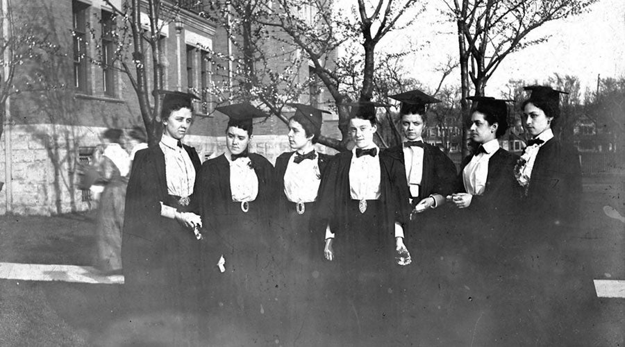 A black and white, faded image of women wearing academic robes standing together talking