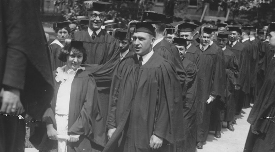 A black and white image of men and women in caps and gowns walking together in a commencement exercise