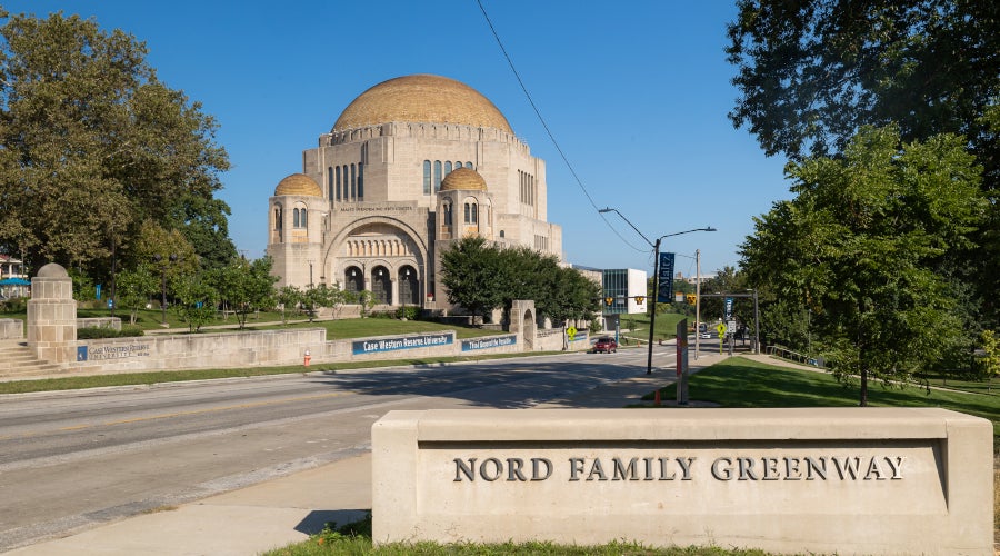  A low-angle shot of the Nord Family Greenway sign in the foreground, looking across a road toward the Maltz Performing Arts Center.
