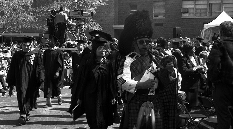 Black and white photo of a piper playing an instrument as he leads graduates gathered behind him across campus