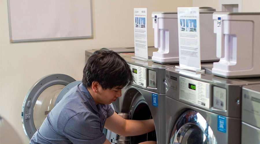A person kneels, inspecting a front-loading washing machine in a brightly lit laundromat. Nearby, detergent dispensers and instruction signs are visible.
