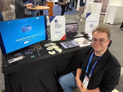 A smiling student sits beside a booth displaying Neura materials at a busy campus showcase.