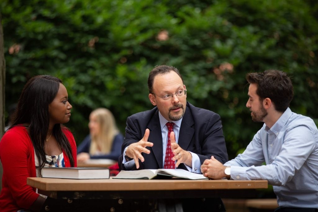Professor Jonathan Adler, with textbooks open, talking with students at the law school on an outdoor picnic table.