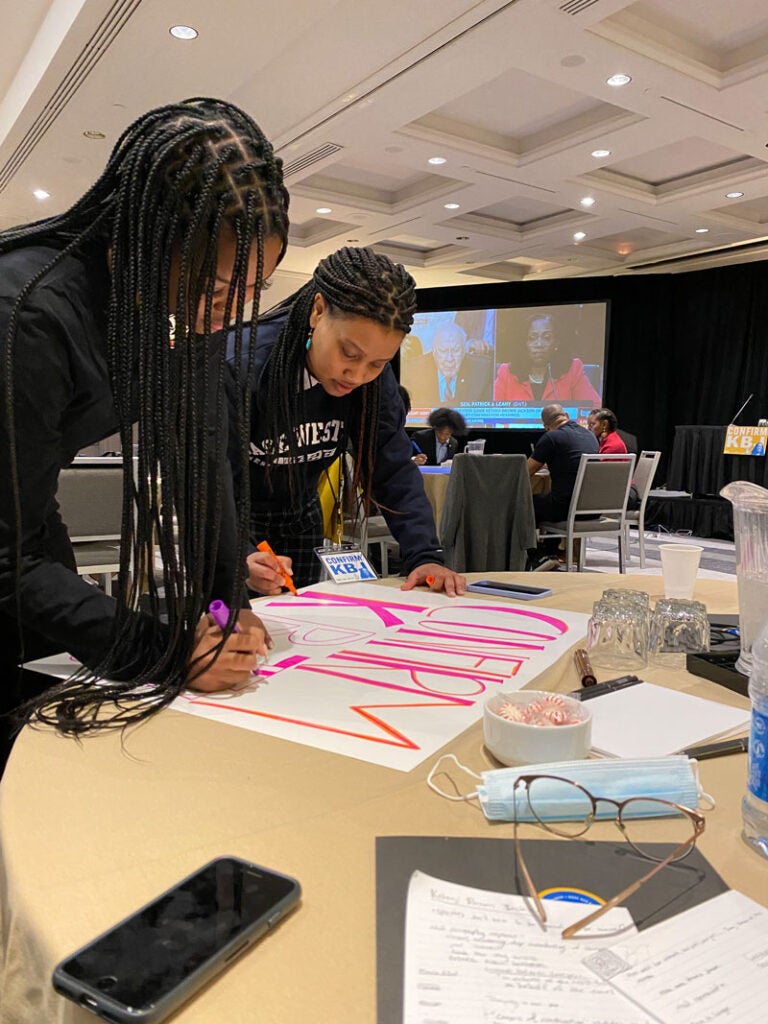 Photo of Case Western Reserve University law student Makela Hayford makes a sign that says "Confirm KBJ" as a senate hearing is on a screen in the background