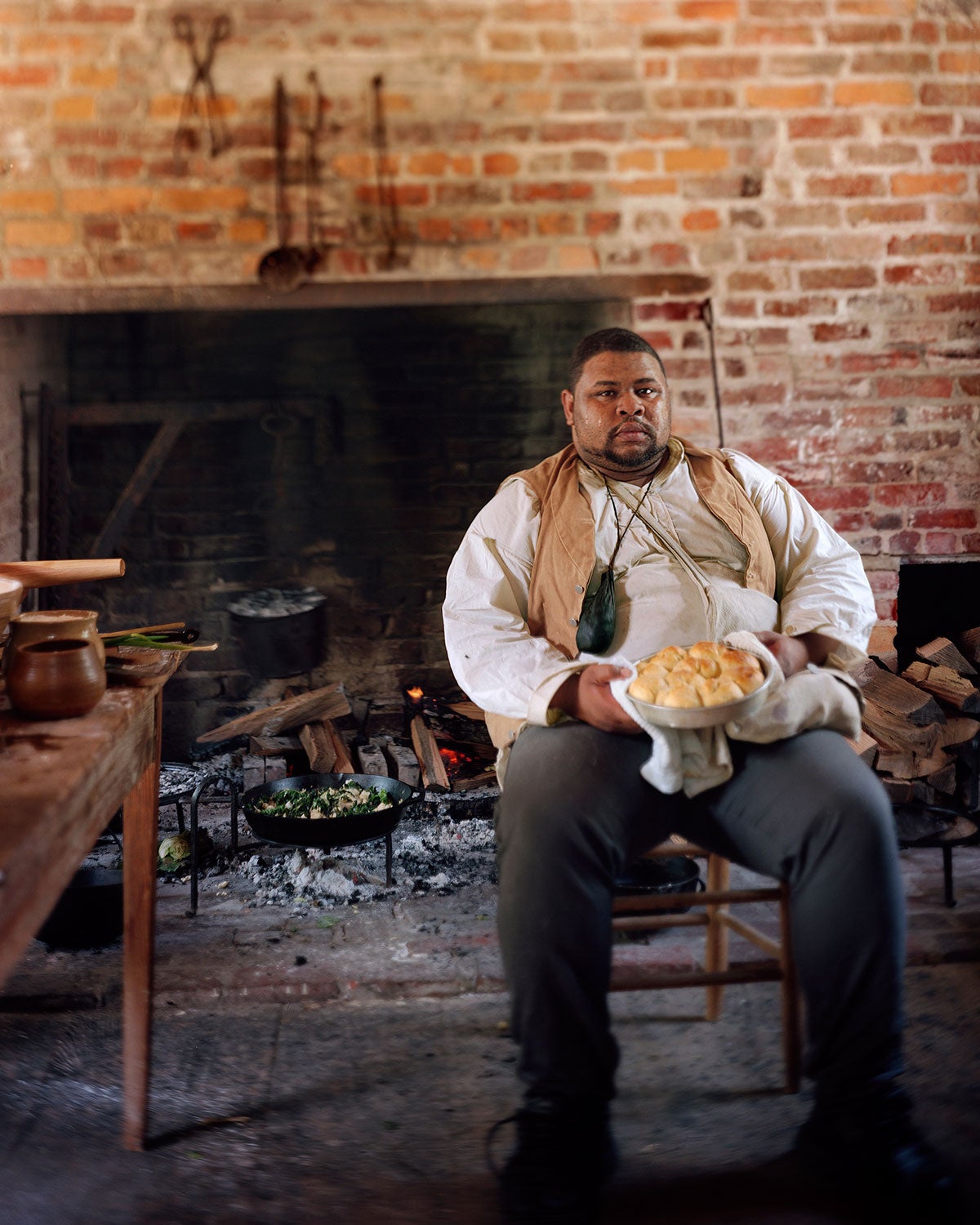 Michael Twitty sitting in front of brick fireplace holding pan of food