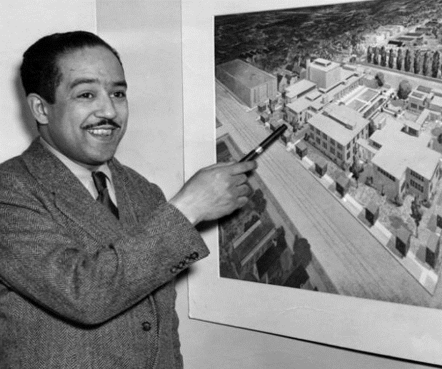 Black and white photo of playwright Langston Hughes pointing to a photo on the wall of buildings in a neighborhood