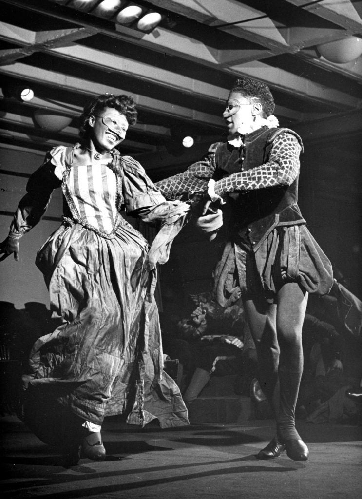 Black and white photo of two costumed actors dancing on stage and wearing masks at Karamu House