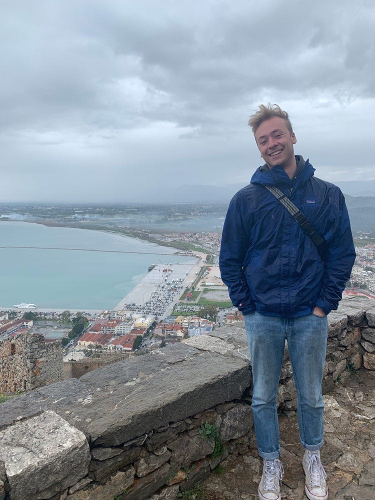 Photo of Colin Hawkinson standing alongside a stone cliff overlooking a sea and city