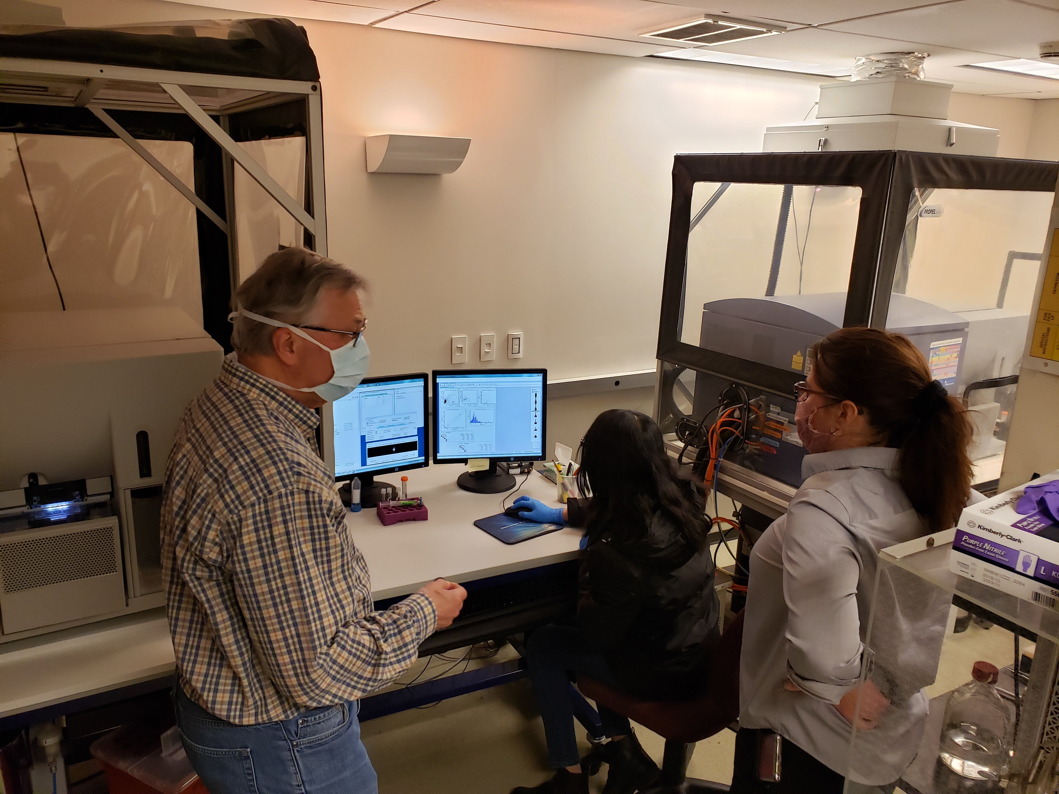 Two people standing in Flow Cytometry Core Facility in front of machines wearing PPE