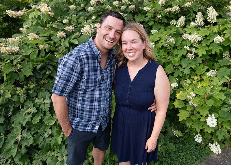 A man in plaid shirt at left with a woman in blue dress at right with green foliage behind them