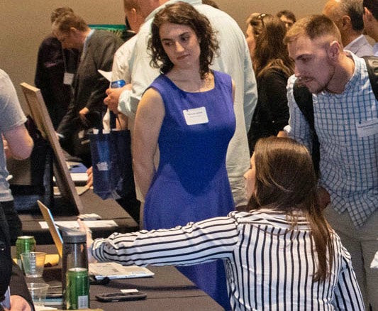 Photo of a woman pointing at something on a computer screen while someone watches on. In the background, three men talk.