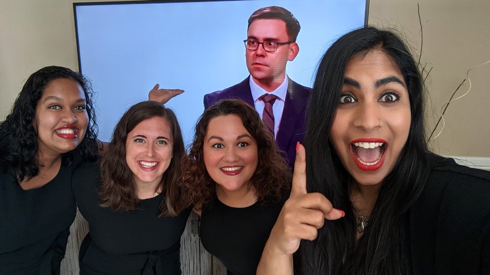 Four alumni pose for a photo with another alum on a TV screen