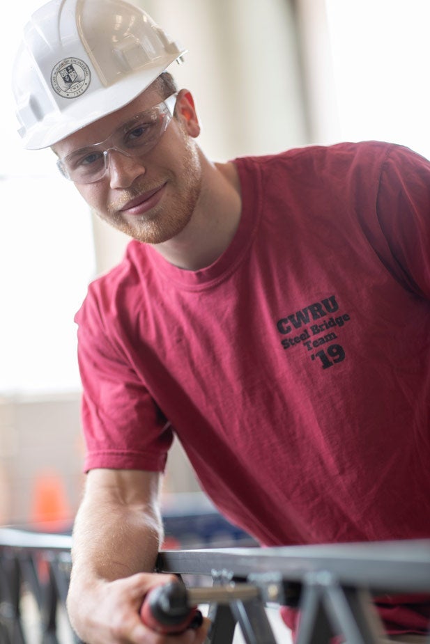 Photo of Luke Traverso holding a tool and wearing a hard hat and safety glasses while leaning on a structure