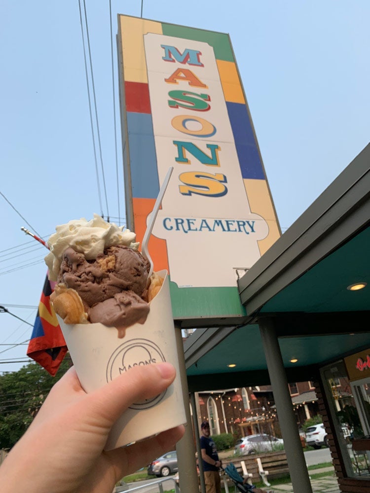 Photo of a hand holding up a cone of Mason's Creamery ice cream in an egg waffle next to the shop's sign