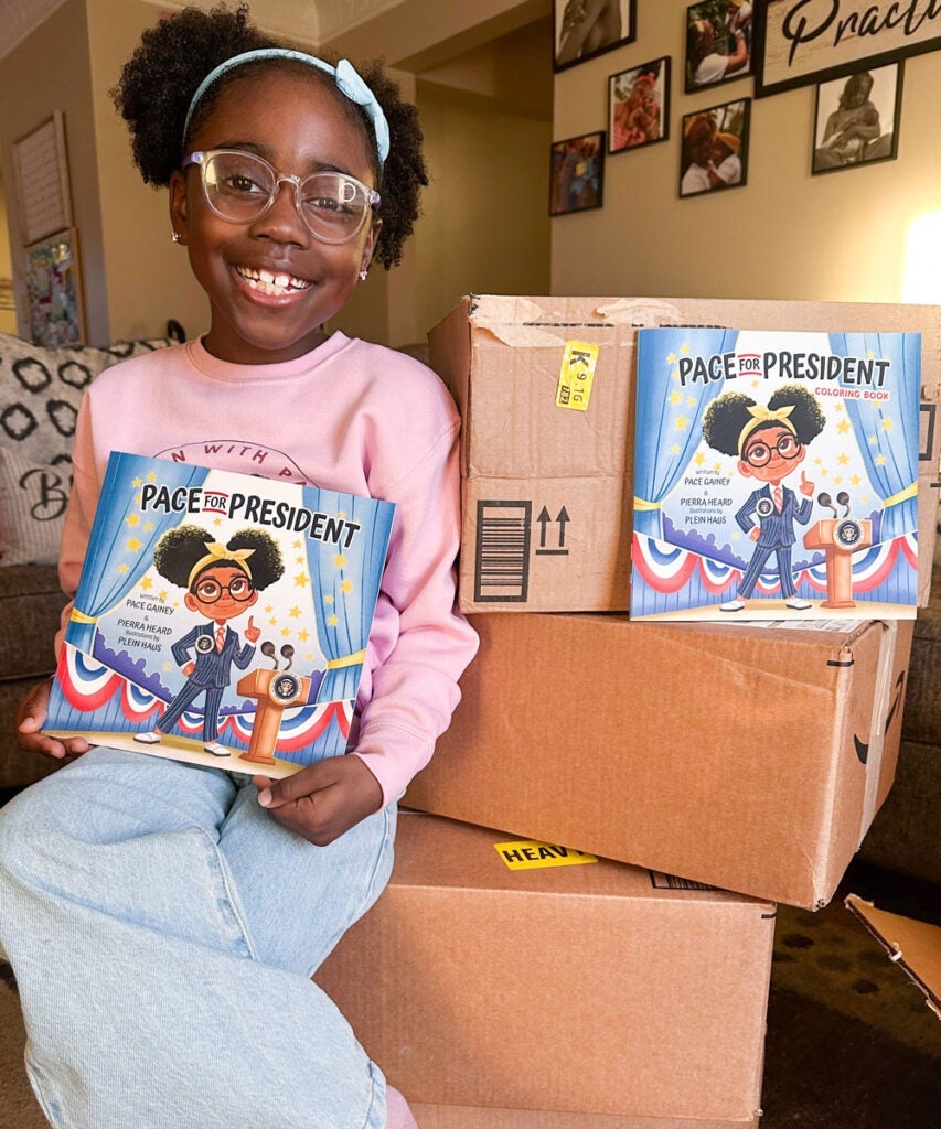 Photo of Pace sitting on boxes of her book, Pace for President, while holding one in her hands