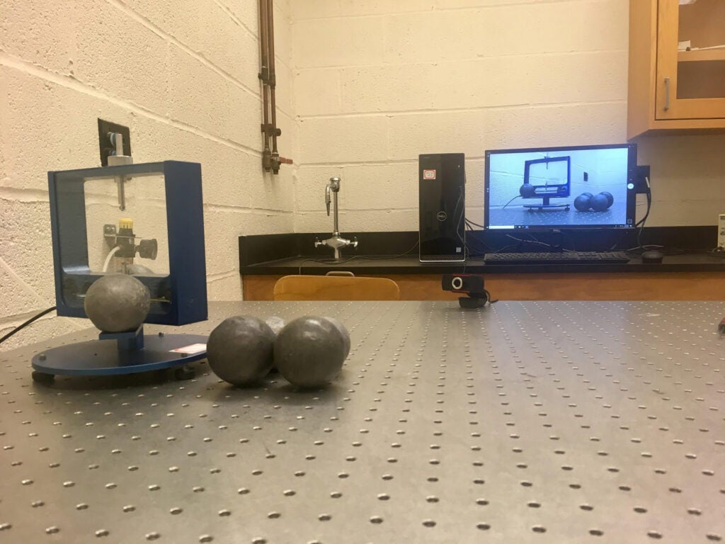 Photo of a lab experiment on a desk with a webcam pointed at it and showing on a computer screen in the background