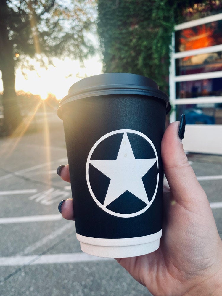 Photo of a hand holding up a Rising Star coffee to-go cup with sun beams shining down