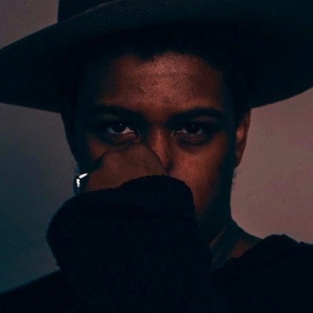 Darkened photo of a man wearing a hat and holding his hand up, obscuring his face