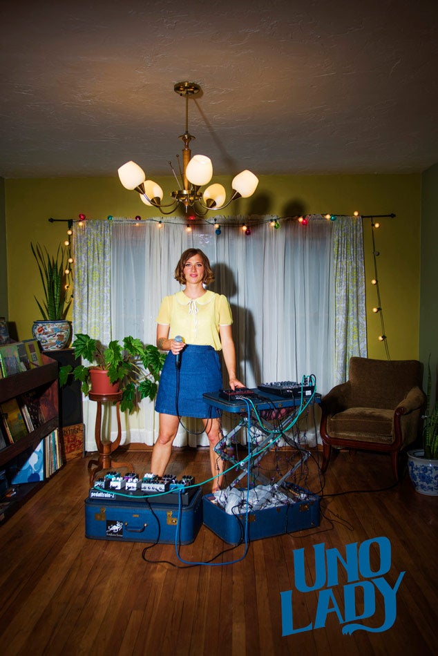 Photo of a woman standing in a living room with a microphone in her hand and sound equipment in front of her and the phrase "Uno Lady" on the bottom of the image