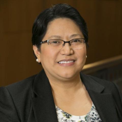 Anna Samia is wearing a black blazer and a patterned blouse, and sitting in front of a wood-paneled background.